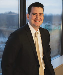Attorney Anthony Gill, of Keches Law Group, is a Caucasian-appearing man with dark hair. He is wearing a white shirt, gold tie, and black suit jacket and is leaning against a window.