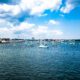 Uma variedade de veleiros ancorados no porto de Boston em meados de junho. O tempo está parcialmente nublado, com céu azul brilhante e algumas nuvens fofas. A água está relativamente calma.