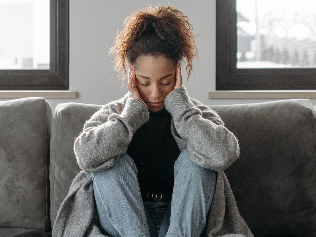 A woman sits on the couch holding her head in pain.